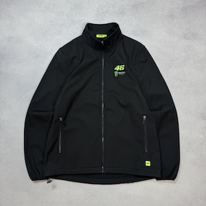 May include: A black softshell jacket featuring a high collar and a full-length front zipper. The chest displays the number 46 in neon yellow above a Monster Energy logo. Two zippered side pockets are present, with a small neon yellow 46 tag.
