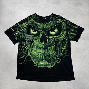 Puede incluir: Camiseta negra con un dise&ntilde;o de calavera verde ne&oacute;n. La calavera tiene ojos brillantes y detalles mec&aacute;nicos. El dise&ntilde;o gr&aacute;fico es detallado y llamativo, cubriendo la parte delantera de la camiseta.