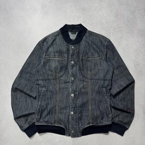 Puede incluir: Chaqueta bomber de mezclilla de lavado oscuro con cierre de botones, dos bolsillos en el pecho y cuello y puños de canalé. La chaqueta presenta costuras visibles y un diseño sutil en la manga.