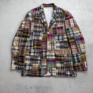 Op de afbeelding: Een patchwork blazer met een veelkleurig ruitpatroon bestaande uit diverse vierkanten van rode, blauwe, gele, groene en witte stof. Het kledingstuk is voorzien van een reverskraag, drie knopen aan de voorzijde, twee klepzakken en vier mouwknopen.