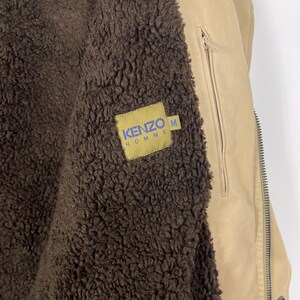 May include: Tan jacket with a brown, fuzzy interior. The jacket has a zipper closure and a small pocket on the chest. A yellow tag with the text "KENZO HOMME" in blue is sewn on the jacket. The jacket is a size M.