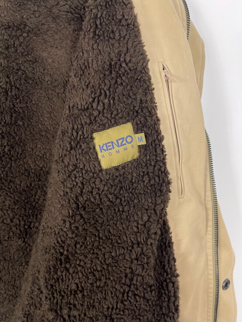May include: Tan jacket with a brown, fuzzy interior. The jacket has a zipper closure and a small pocket on the chest. A yellow tag with the text "KENZO HOMME" in blue is sewn on the jacket. The jacket is a size M.