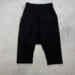 May include: Black cropped trousers with two front pockets and a button closure. The trousers have a relaxed fit and an asymmetrical design. The fabric appears to be a medium-weight material, suitable for casual wear.
