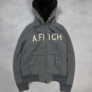 May include: A grey zip-up hoodie with a fur-lined hood. The hoodie has the text "A. Fitch" across the chest in a cream-coloured, textured font. It features two front pockets and ribbed cuffs and hem.