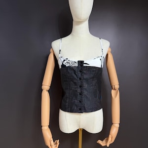 May include: A sleeveless corset-style top featuring a black lower section with a vertical row of buttons and a white upper section with a black floral pattern. The garment has thin white spaghetti straps and a straight neckline, displayed on a mannequin.