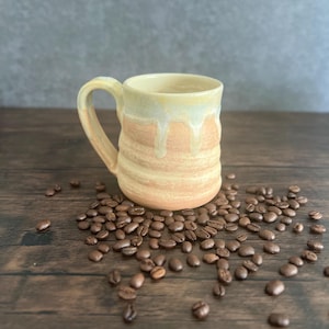 May include: A handmade ceramic mug with a dripped glaze effect. The mug features a beige and orange color scheme, with a textured surface. Coffee beans surround the mug on a wooden surface.