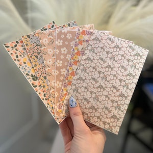 May include: A hand displays a selection of floral patterned cards. The cards showcase diverse designs with small flowers in pink, yellow, orange, and white against neutral backgrounds. The rectangular cards are likely stationery or paper goods.