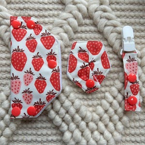Peut inclure: Un ensemble de trois accessoires en tissu avec un motif de fraises rouges sur fond blanc. Le lot comprend une enveloppe rectangulaire avec quatre boutons-pression rouges, une petite pièce pentagonale avec un bouton-pression et une sangle à pince.