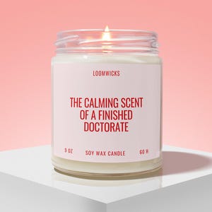 May include: A clear glass jar soy wax candle with a lit wick. The white label features the text LOOMWICKS, THE CALMING SCENT OF A FINISHED DOCTORATE, 9 OZ, and 60 H in red lettering. The candle is filled with white wax and has a visible flame.