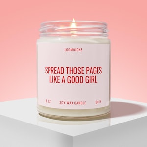 May include: A lit soy wax candle in a clear glass jar with a white label. The label features the text LOOMWICKS, SPREAD THOSE PAGES LIKE A GOOD GIRL, 9 OZ, and 60 H in red lettering. The candle has a visible flame and is filled with white wax.