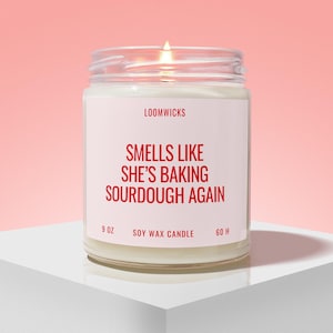 May include: A lit soy wax candle in a clear glass jar with a white label. The label features the text LOOMWICKS, SMELLS LIKE SHE'S BAKING SOURDOUGH AGAIN, 9 OZ, SOY WAX CANDLE, and 60 H in red lettering against a light pink background.