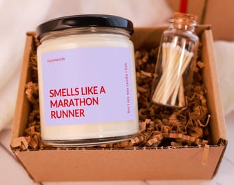 May include: A clear glass jar candle with a black lid and a light purple label featuring the red text "SMELLS LIKE A MARATHON RUNNER" and "9oz | soy wax candle | 60h". Next to it is a small glass bottle filled with wooden matches inside a box.