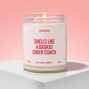 May include: A clear glass jar candle with a white label featuring the text LOOMWICKS, SMELLS LIKE A BADASS CHEER COACH, 9 OZ, SOY WAX CANDLE, and 60 H. The candle contains white wax and has a lit wick, emitting a small flame from the center.