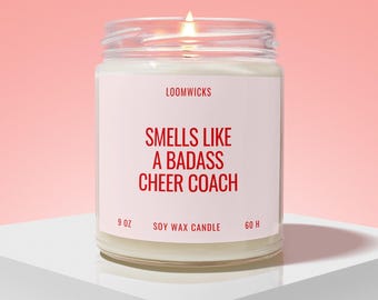 May include: A clear glass jar candle with a white label featuring the text LOOMWICKS, SMELLS LIKE A BADASS CHEER COACH, 9 OZ, SOY WAX CANDLE, and 60 H. The candle contains white wax and has a lit wick, emitting a small flame from the center.