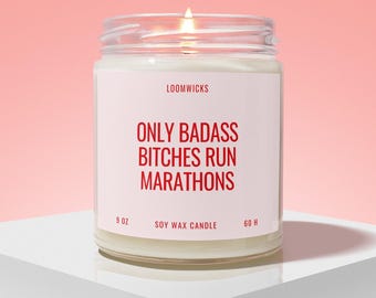 May include: A lit soy wax candle in a clear glass jar with a white label. The label features the red text LOOMWICKS, ONLY BADASS BITCHES RUN MARATHONS, 9 OZ, SOY WAX CANDLE, and 60 H. The candle has a visible flame and white wax inside the glass container.