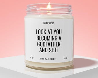 May include: A lit soy wax candle in a clear glass jar with a white label. The label features the text LOOMWICKS and LOOK AT YOU BECOMING A GODFATHER AND SHIT, along with 9 OZ and 60 H printed at the bottom. The candle has a visible flame at the top.