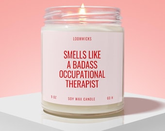 May include: A clear glass jar candle with a white label featuring red text that reads LOOMWICKS, SMELLS LIKE A BADASSS OCCUPATIONAL THERAPIST, 9 OZ, SOY WAX CANDLE, and 60 H. The candle has white wax and a lit wick, emitting a warm glow from the center.