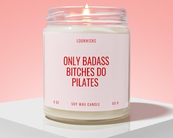 May include: A clear glass jar candle with a white label featuring the red text LOOMWICKS, ONLY BADASS BITCHES DO PILATES, 9 OZ, SOY WAX CANDLE, and 60 H. The candle has white wax and a lit wick, creating a warm glow against a soft pink background.