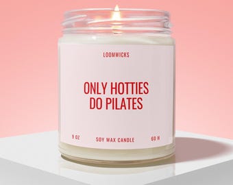 May include: A clear glass jar candle with a white label featuring the red text LOOMWICKS, ONLY HOTTIES DO PILATES, 9 OZ, SOY WAX CANDLE, and 60 H. The candle has white wax and a lit wick, emitting a small flame from the center of the jar.