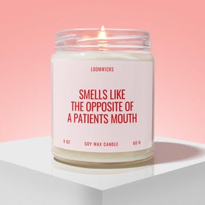 May include: A lit soy wax candle in a clear glass jar with a white label. The label features the text LOOMWICKS, 9 OZ, 60 H, and the red printed phrase SMELLS LIKE THE OPPOSITE OF A PATIENTS MOUTH. The candle wax is white and the flame is visible.