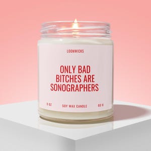 May include: A clear glass jar soy wax candle with a lit wick. The white label features the red text LOOMWICKS, ONLY BAD BITCHES ARE SONOGRAPHERS, 9 OZ, SOY WAX CANDLE, and 60 H. The candle is filled with white wax and has a visible flame at the top.