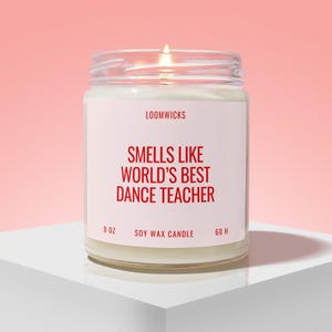 May include: A clear glass jar candle with a white label featuring the red text LOOMWICKS, SMELLS LIKE WORLD'S BEST DANCE TEACHER, 9 OZ, SOY WAX CANDLE, and 60 H. The candle contains white wax and has a lit wick visible at the top of the glass container.