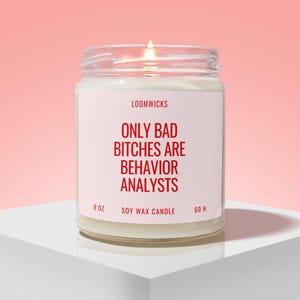 May include: A lit soy wax candle in a clear glass jar with a white label featuring the red text LOOMWICKS, ONLY BAD BITCHES ARE BEHAVIOR ANALYSTS, 9 OZ, and 60 H. The candle has a visible flame and is filled with white wax, presented in a simple container.