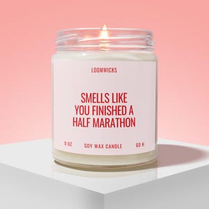 May include: A clear glass jar candle with a white label featuring red text that reads LOOMWICKS, SMELLS LIKE YOU FINISHED A HALF MARATHON, 9 OZ, SOY WAX CANDLE, and 60 H. The candle has a lit wick and contains white wax.