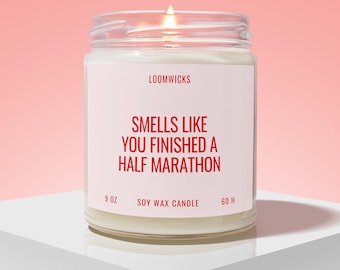 May include: A clear glass jar candle with a white label featuring red text that reads LOOMWICKS, SMELLS LIKE YOU FINISHED A HALF MARATHON, 9 OZ, SOY WAX CANDLE, and 60 H. The candle has a lit wick and contains white wax.