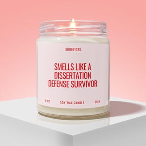 May include: A clear glass jar candle with a white label featuring the red text LOOMWICKS, SMELLS LIKE A DISSERTATION DEFENSE SURVIVOR, 9 OZ, SOY WAX CANDLE, and 60 H. The candle has a lit wick and is filled with white wax, showing a warm glow.