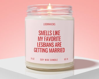 Peut inclure: Une bougie en cire de soja allumée dans un pot en verre transparent avec une étiquette blanche portant le texte LOOMWICKS, SMELLS LIKE MY FAVORITE LESBIANS ARE GETTING MARRIED, 9 OZ, 60 H. La bougie a une surface en cire blanche et une flamme.