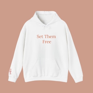 May include: White hoodie with a front pocket and drawstring hood. The phrase "Set Them Free" is printed in a reddish-brown color across the chest. A small graphic is on the left sleeve. The hoodie is made of a soft material.