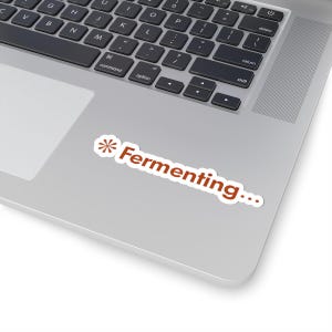 May include: A white sticker with the text "Fermenting..." in a reddish-brown font, with a stylized asterisk symbol at the beginning. The sticker is on a silver laptop.