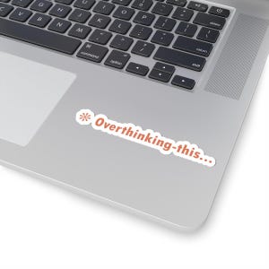 May include: A white sticker with the text "Overthinking-this..." in orange, placed on a silver laptop. The sticker has a small orange asterisk design on the left side.