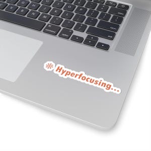 May include: A white sticker with the text "Hyperfocusing..." in orange, and a small orange floral design. The sticker is on a silver laptop, near the keyboard.