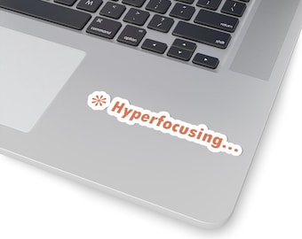 Puede incluir: Una pegatina blanca troquelada que presenta el texto "Hyperfocusing..." en una fuente sans-serif en negrita de color terracota, acompañada de un pequeño asterisco a juego del mismo color a la izquierda de la palabra, aplicada sobre un portátil plateado.