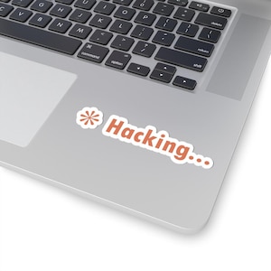 May include: A white sticker with the orange text "Hacking..." on a laptop. The sticker features a flower design. The laptop's keyboard is visible in the background. The laptop is silver.