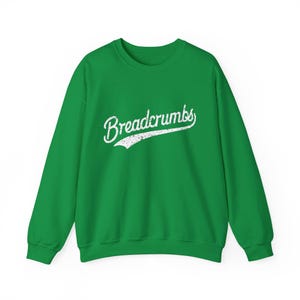 May include: A green sweatshirt featuring the word "Breadcrumbs" in a white, cursive font. The text has a distressed, vintage appearance. The sweatshirt is a crew neck style with long sleeves.