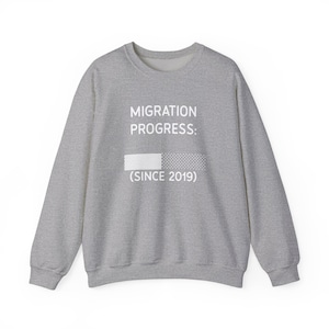 Pode incluir: Uma camisola de gola redonda cinzenta mesclada com o texto branco MIGRATION PROGRESS: seguido de uma barra de carregamento gráfica e o texto (SINCE 2019) centrado no peito. A peça tem mangas compridas e uma gola canelada.