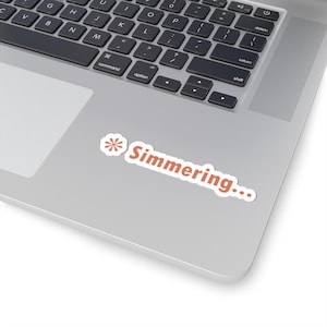 May include: A white sticker with the word "Simmering..." in orange script. The sticker has a small orange and white floral design on the left side. The sticker is on a silver laptop.