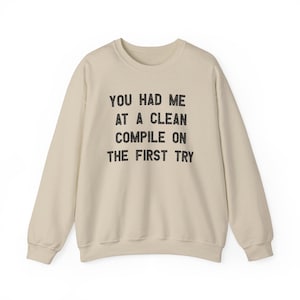 Puede incluir: Sudadera de color crema con el texto negro "YOU HAD ME AT A CLEAN COMPILE ON THE FIRST TRY". La sudadera tiene cuello redondo y mangas largas, ideal para uso casual.