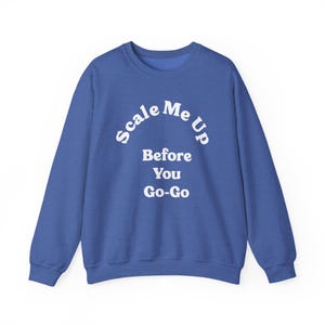 May include: A royal blue crewneck sweatshirt featuring the white text Scale Me Up Before You Go-Go printed in a bold, sans-serif font across the chest. The garment has long sleeves, a ribbed neckline, and ribbed cuffs, providing a standard fit.