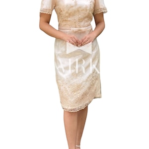 May include: A cream-coloured, two-piece dress with intricate floral embroidery. The top features a square neckline and short, puffed sleeves. The skirt is knee-length. The dress is accessorised with a matching bow at the waist and white sandals.