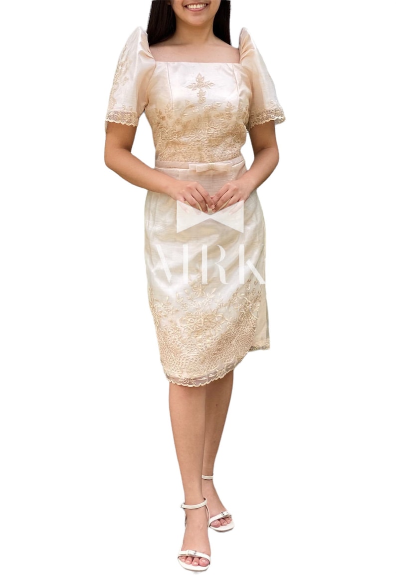 May include: A cream-coloured, two-piece dress with intricate floral embroidery. The top features a square neckline and short, puffed sleeves. The skirt is knee-length. The dress is accessorised with a matching bow at the waist and white sandals.