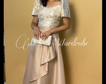 May include: A white and gold floral patterned dress with a ruffled skirt. The dress has a scoop neckline and short sleeves. The model is holding a white clutch.