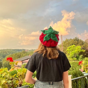May include: A person wearing a red and green knitted strawberry hat, a black t-shirt, and light blue denim shorts. The person is standing on a balcony overlooking a green hillside with a cloudy sky in the background.