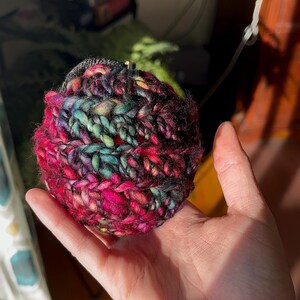 Pu&ograve; includere: Una mano che tiene una palla di lana colorata con lana rosa, nera, verde e blu. La palla di lana &egrave; all'uncinetto e ha una superficie testurizzata.