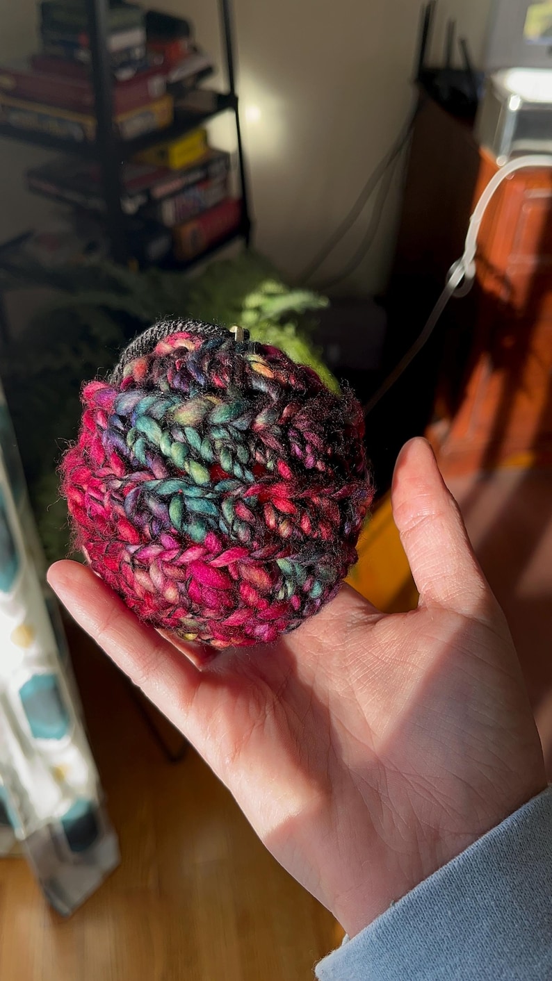 Pu&ograve; includere: Una mano che tiene una palla di lana colorata con lana rosa, nera, verde e blu. La palla di lana &egrave; all'uncinetto e ha una superficie testurizzata.