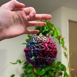 Pu&ograve; includere: Un ornamento natalizio colorato, lavorato a maglia a mano, in tonalit&agrave; di rosa, viola, blu e verde. L'ornamento &egrave; rotondo e ha una superficie testurizzata.