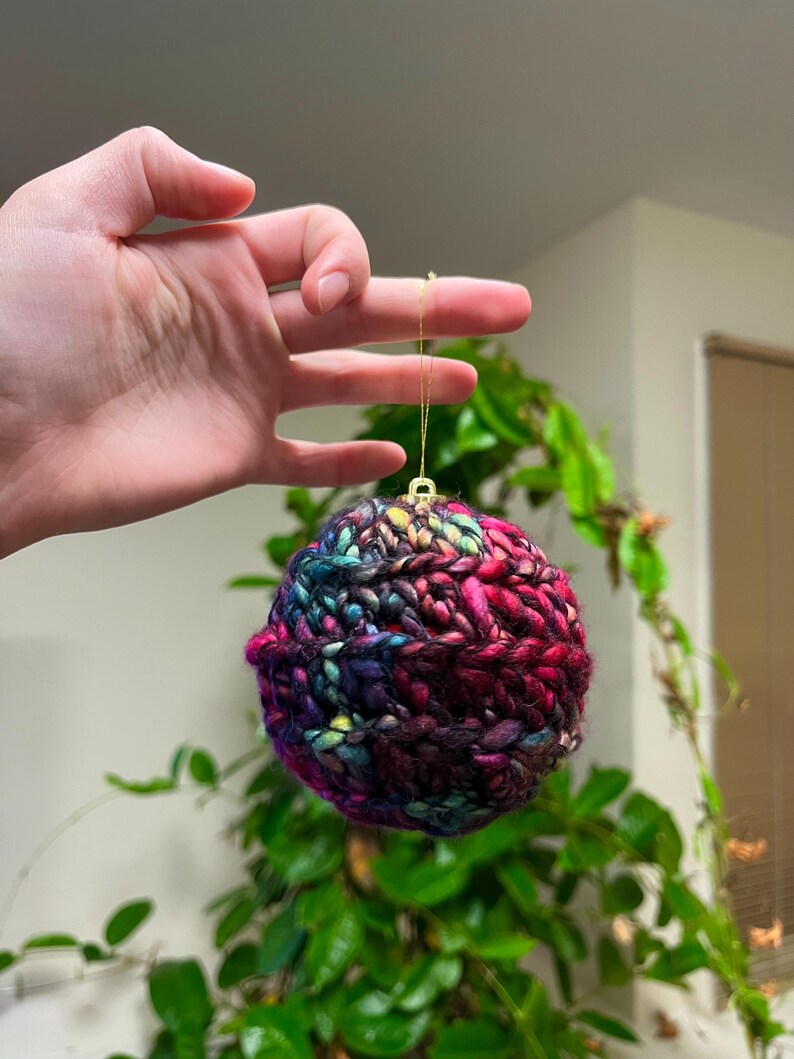 Pu&ograve; includere: Un ornamento natalizio colorato, lavorato a maglia a mano, in tonalit&agrave; di rosa, viola, blu e verde. L'ornamento &egrave; rotondo e ha una superficie testurizzata.